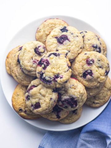 A batch of blueberry cookies on a round white plate placed on top of a blue napkin to the bottom right of the plate going out of frame.