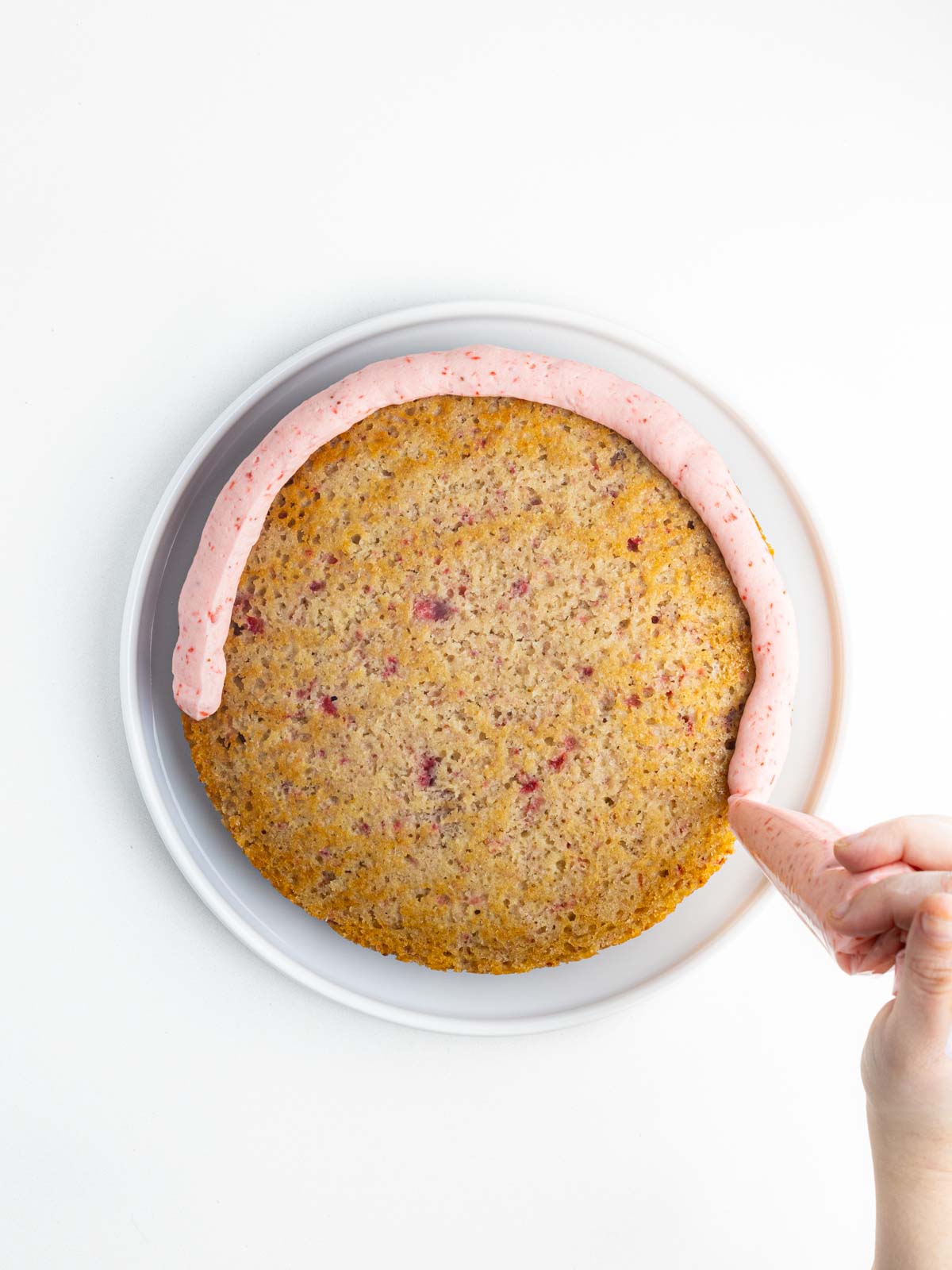 A hand halfway through piping a circle of pink frosting around the outside edge of the cake.