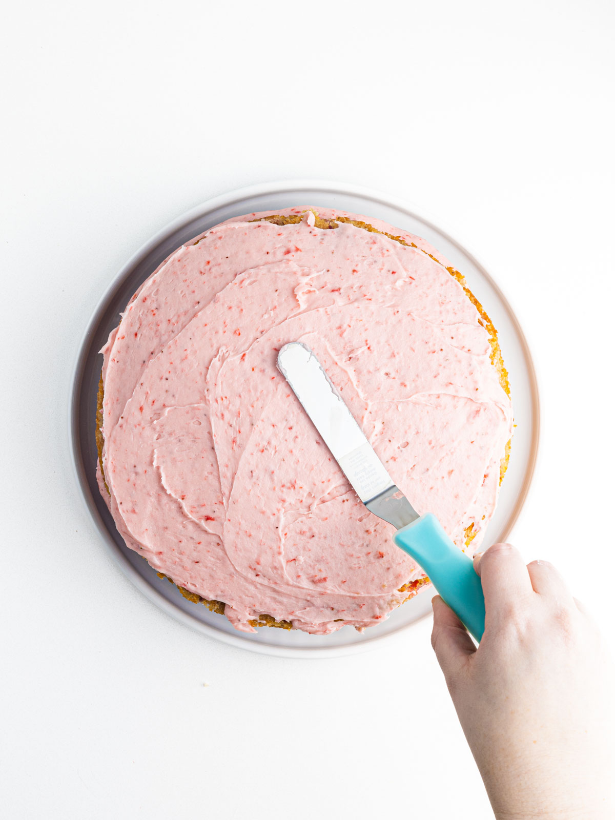 A hand holding an offset spatula with a blue handle spreading pink frosting over the top of the cake.