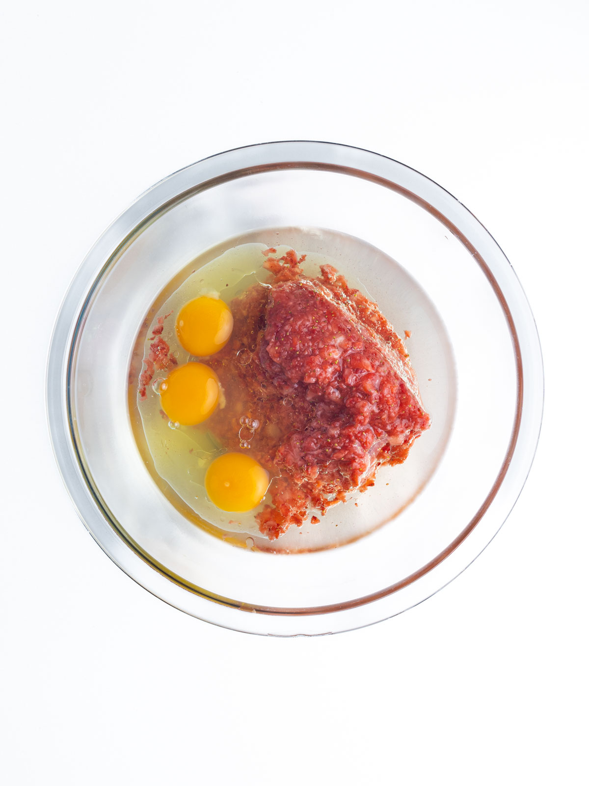 Strawberry puree, oil, and eggs in a clear glass mixing bowl on a white surface.