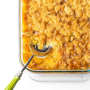 The bottom left corner of a 9x13 baking dish of peach cobbler with a serving spoon with a green handle sticking out of the dish.