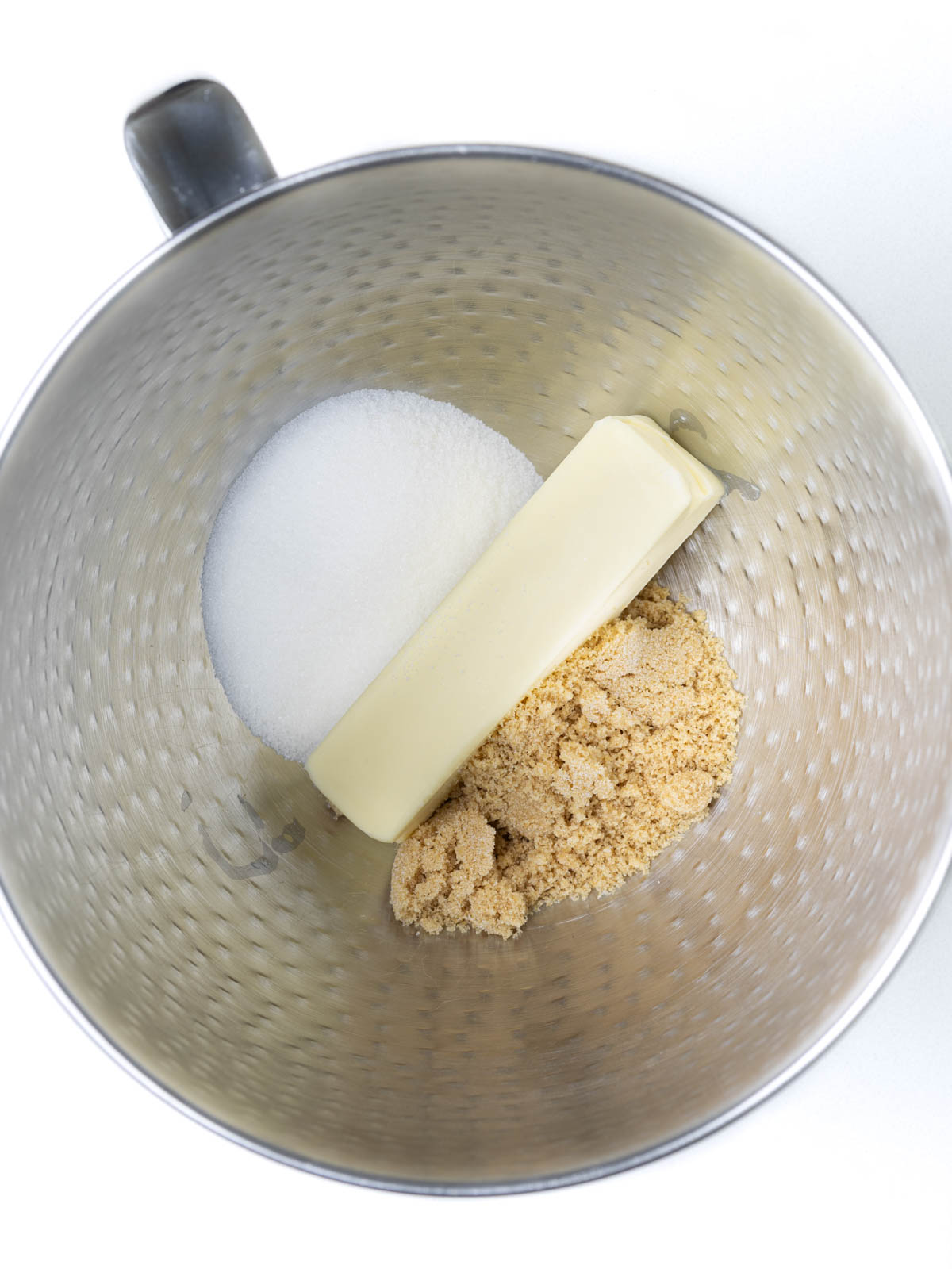 Butter, sugar, and brown sugar in a textured stainless steel mixing bowl with a handle.